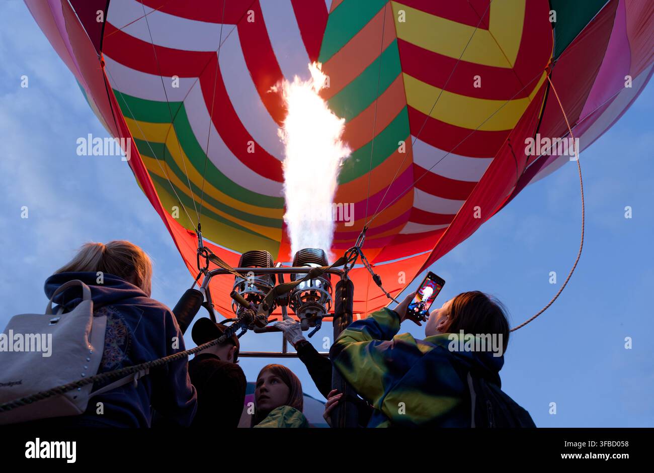 Niedrigwinklige Aufnahme von Menschen, die zuschauen und Fotos machen, während eine starke Propaneflamme einen farbenfrohen Heißluftballon entzündet. Stockfoto