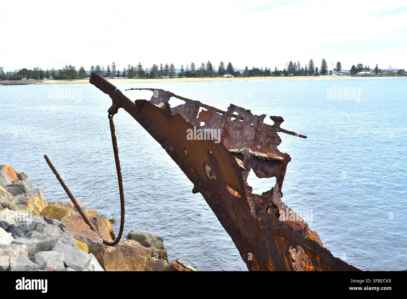 Ein gezacktes, verrostetes Wrack ragt über ruhiges Küstewasser hinaus, sein korrodiertes Metallgerüst ist von großen Löchern und gebrochenen Kanten gekennzeichnet. Stockfoto