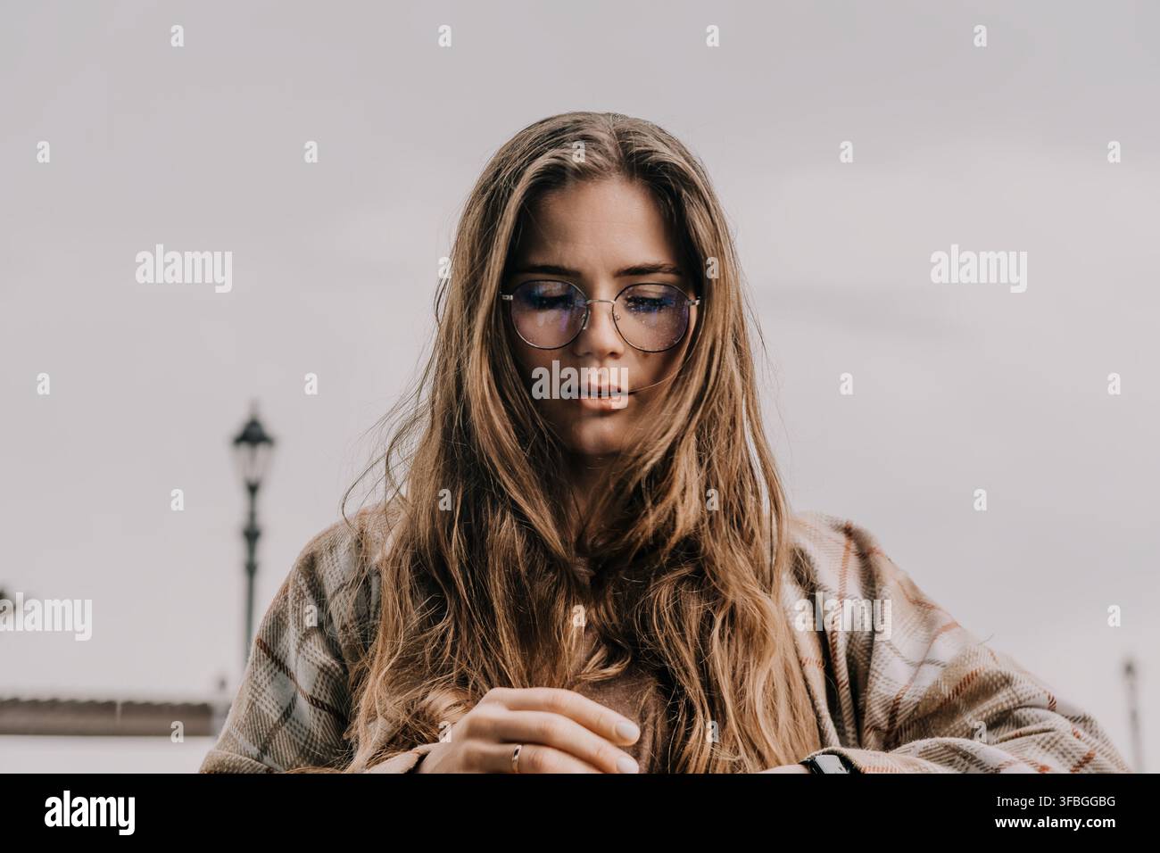 Frau mit Brille, junges Mädchen in stilvollen Brillen und kariertem Mantel schaut nach unten im Freien unter einem bewölkten Himmel Stockfoto