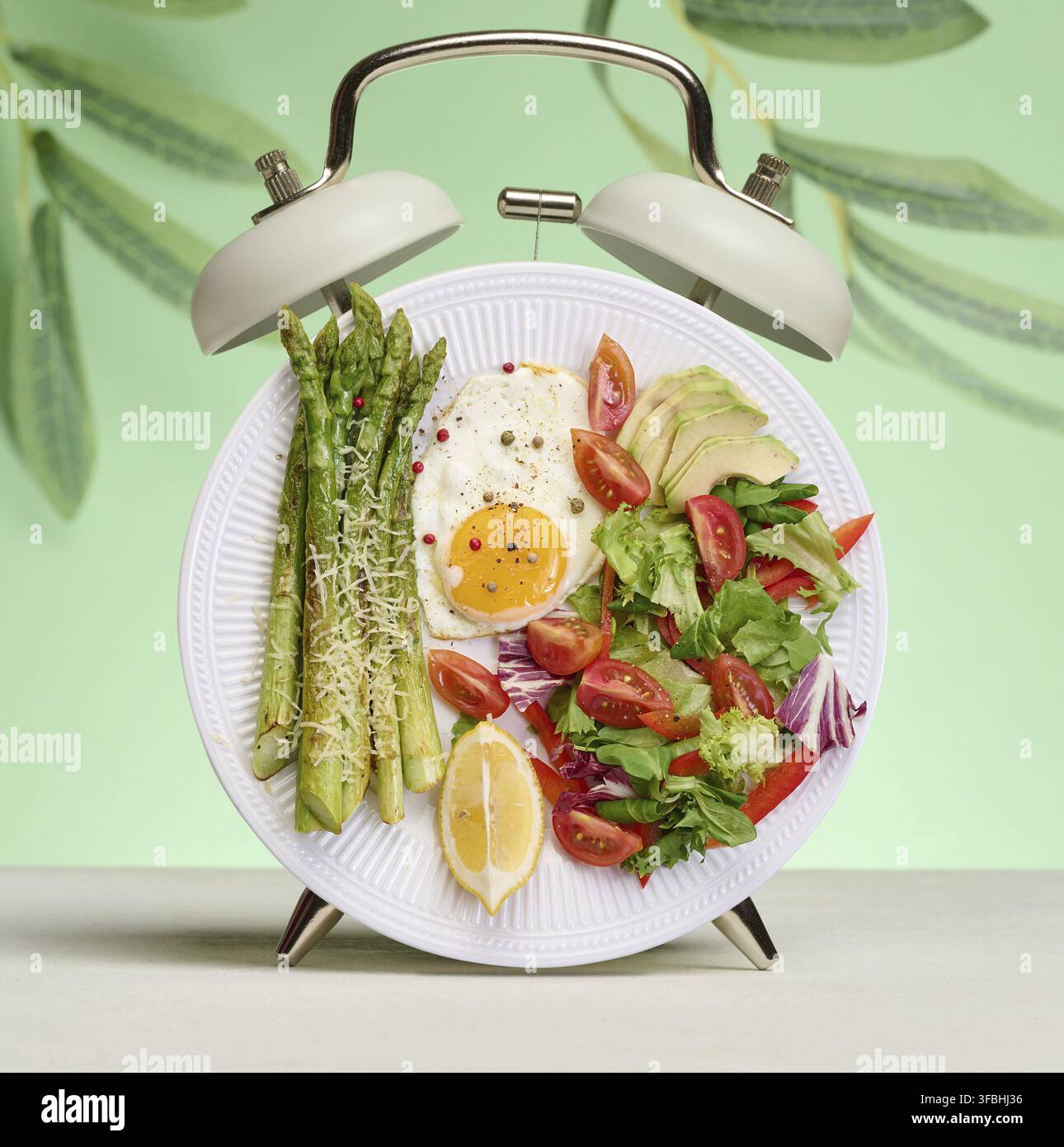 Teller mit Spargel, Spiegeleiern und Gemüsesalat, stilisiert als Wecker auf grünem Hintergrund. Gesunde Lebensweise und Ernährung Stockfoto