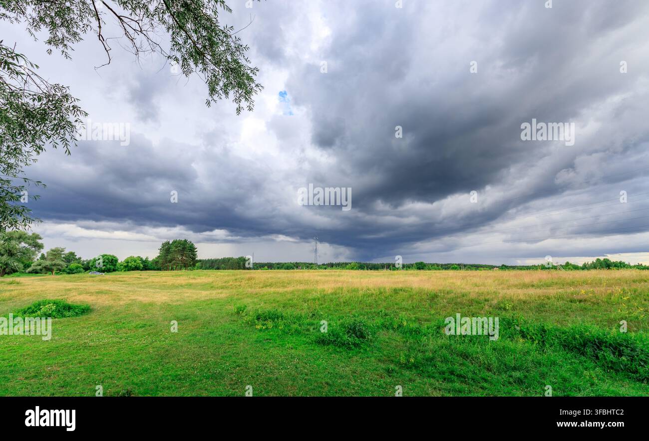 Dunkle Sturmwolken türmen sich über einem weiten grünen Feld. Bäume rahmen die Szene links und rechts ein Stockfoto