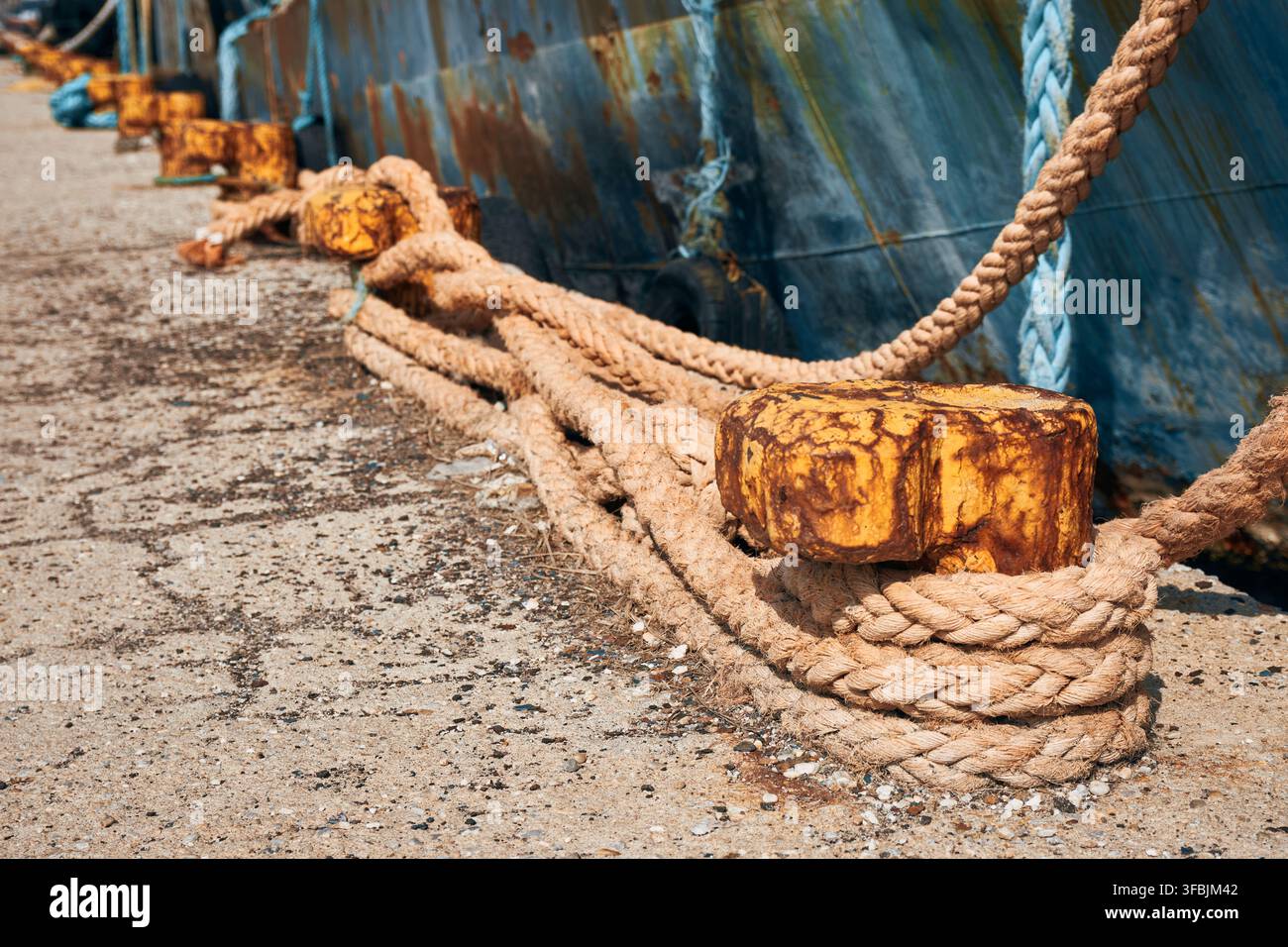 Nahaufnahme-Perspektive von verwitterten, rostigen Metall-Befestigungspfeilern und schweren geflochtenen Hanfseilen, die sicher an einem Betonpier befestigt sind und ein kommerzielles Schiff halten Stockfoto