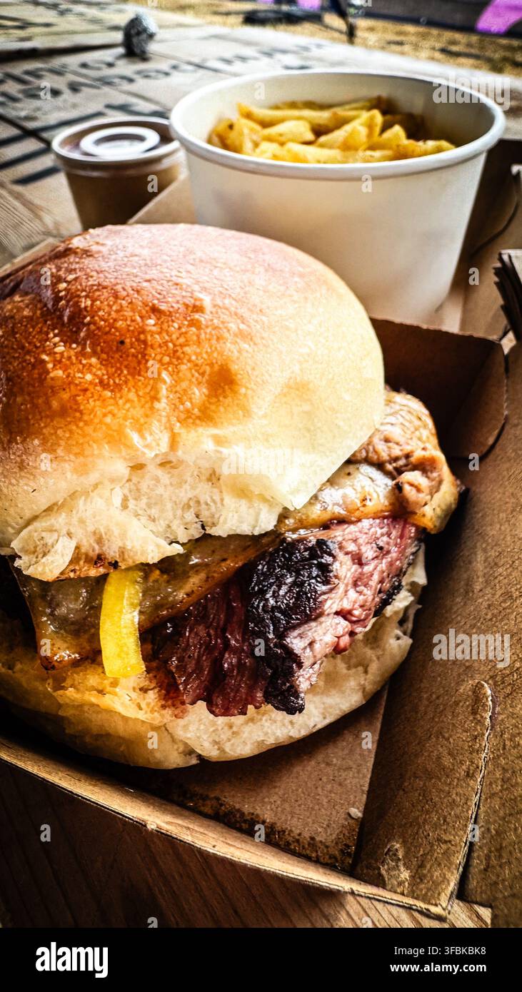 Gourmet-Steakburger mit geschmolzenem Käse und Gurken, serviert mit Pommes und einem Getränk in einer Takeaway-Box von einem Imbisswagen - Smartphone-aufgenommenes Stockfoto