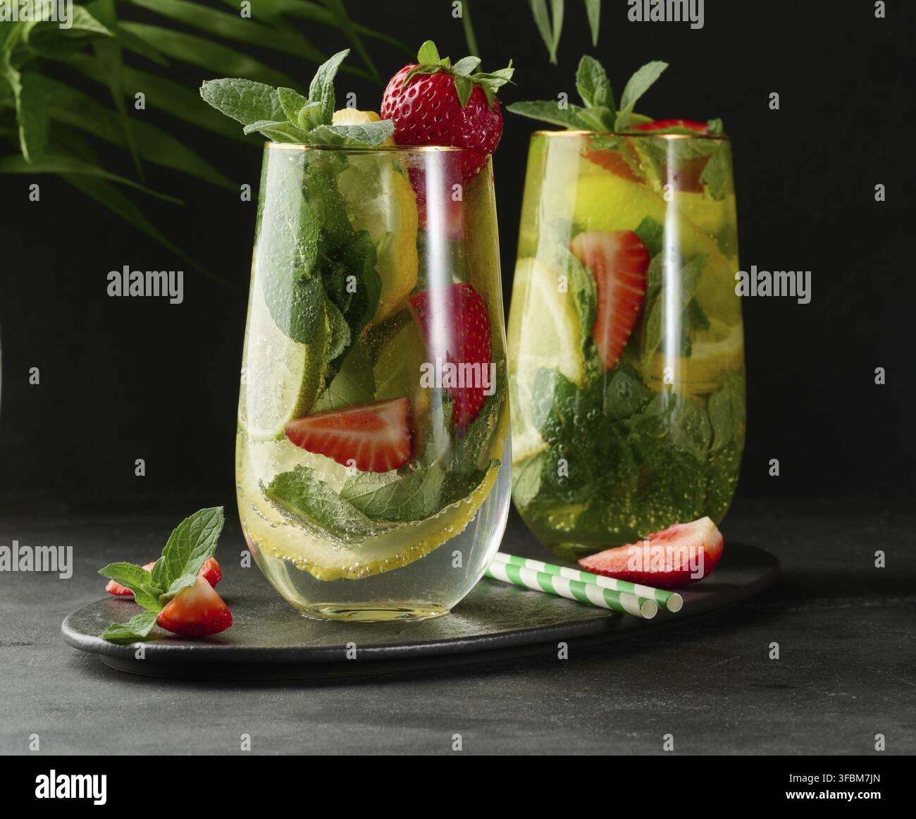 Ein erfrischendes Getränk im Glas mit Zitronenscheiben, Erdbeeren, frischen Minzblättern und Mineralwasser, serviert mit Limette und Minze auf einem schwarzen Teller Stockfoto