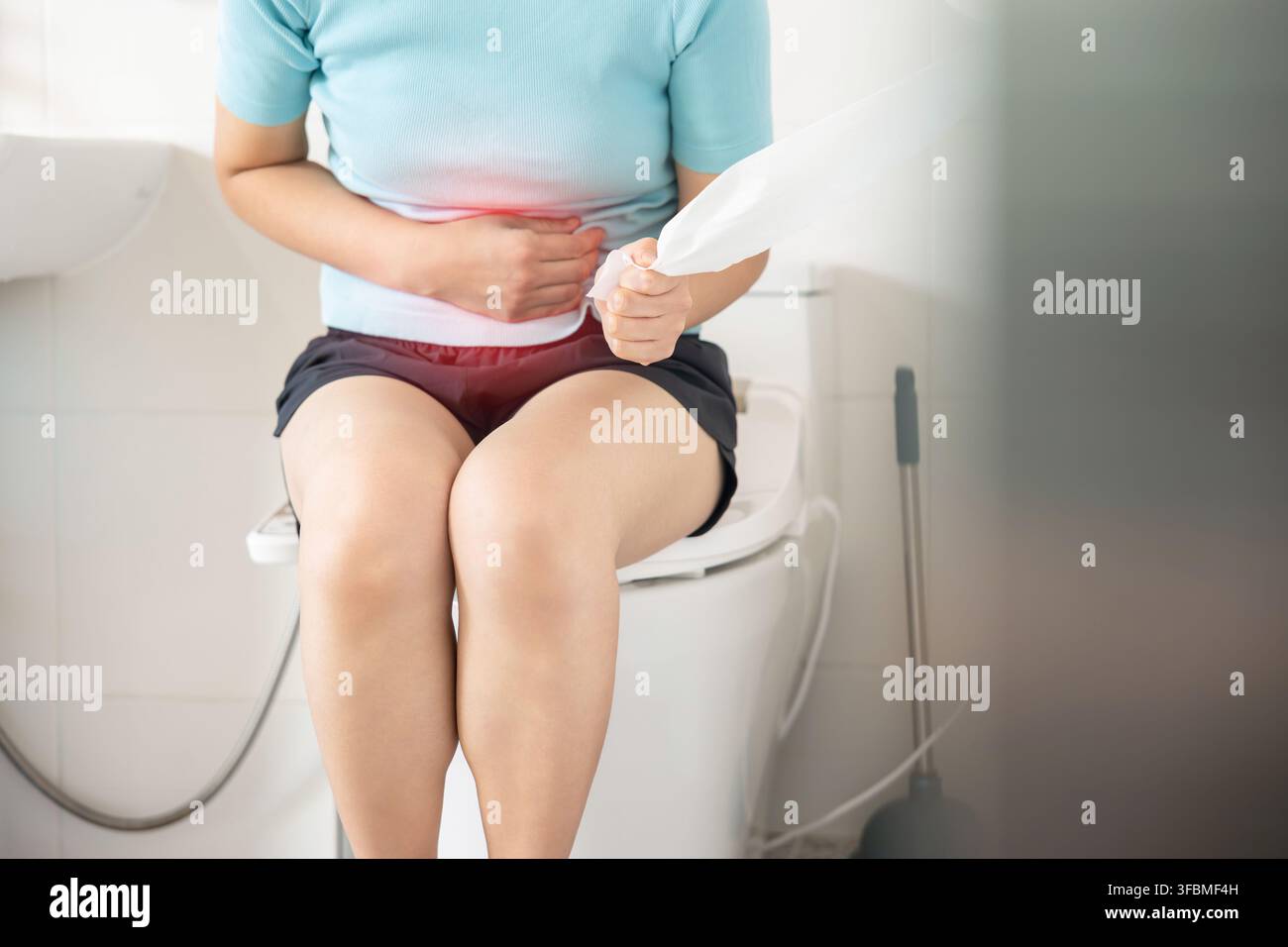 Koreanerin auf einer Toilette, die Toilettenpapier hält, mit einer roten visuellen Markierung, die auf Bauchschmerzen und Verstopfung hinweist. Stockfoto