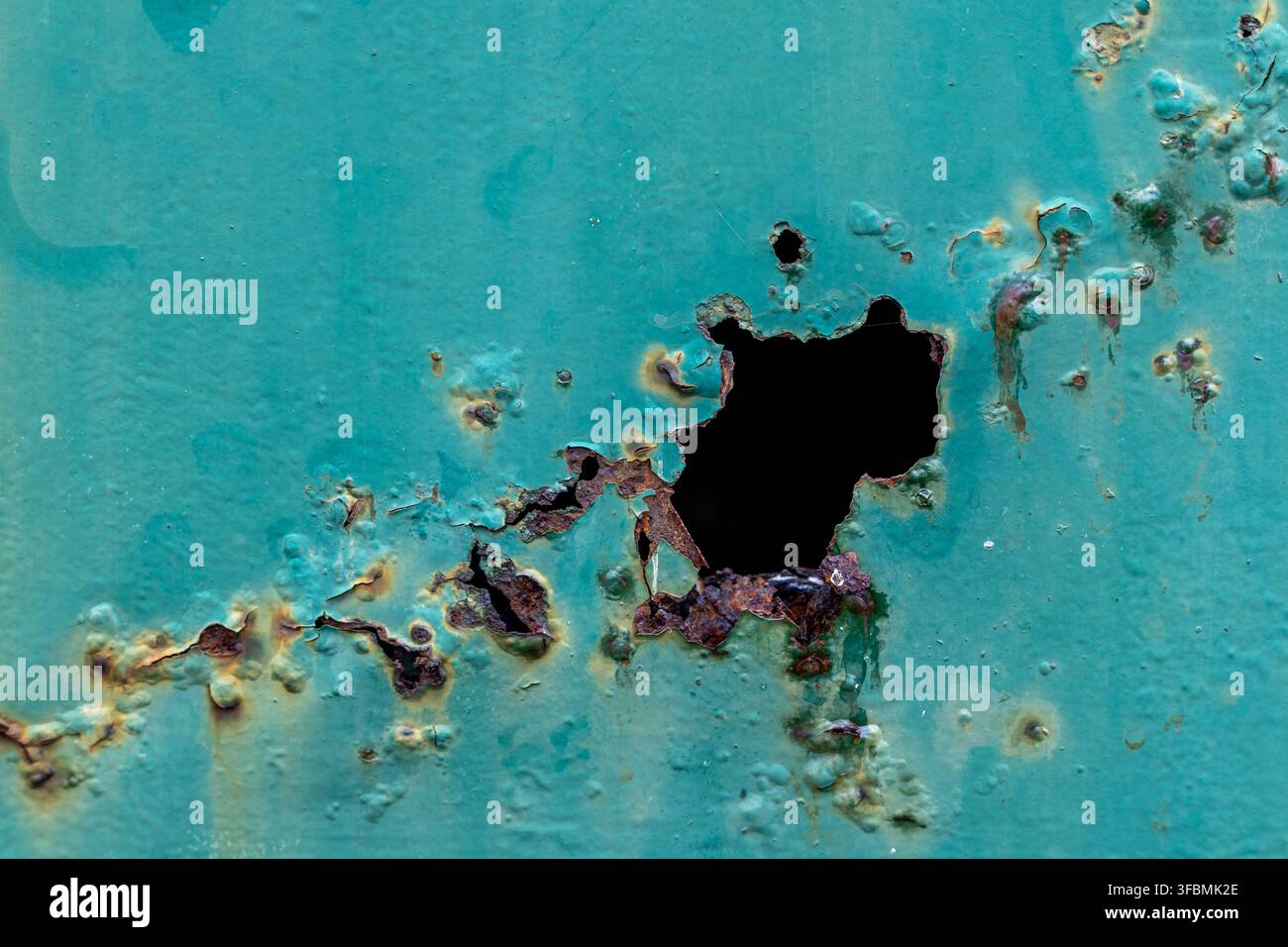 Korrodiertes Metalloberfläche mit einem großen Rostloch. Abblätternde türkisfarbene Farbe umgibt den beschädigten Bereich. Kleinere Rostflecken und Lochbildung. Stockfoto