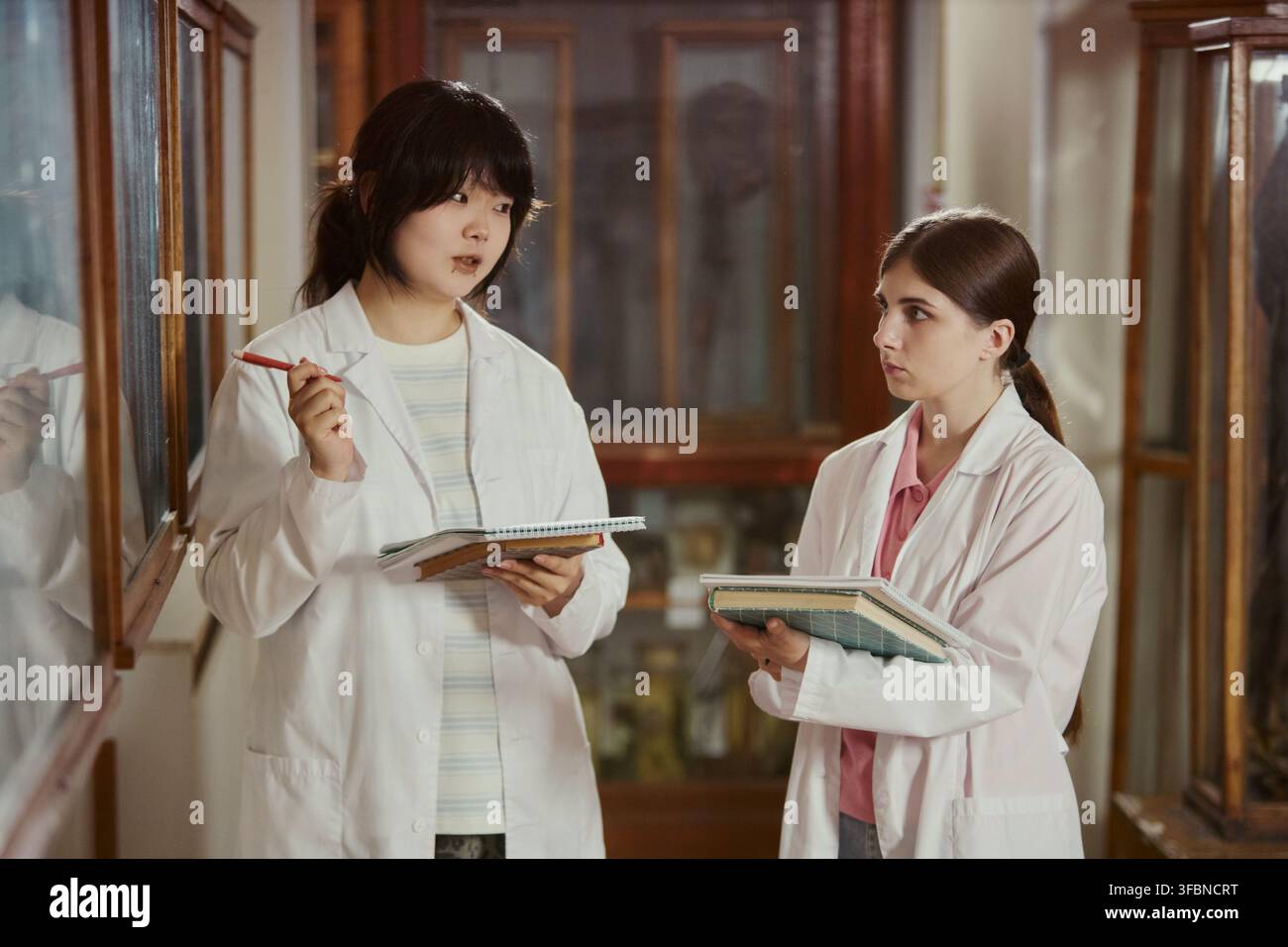 Asiatische junge Erwachsene und kaukasische junge erwachsene Studentinnen tragen Laborkittel, halten Notizbücher und diskutieren Forschung im Museum. Bild geeignet für die Universitäten-Marketingbildung Stockfoto