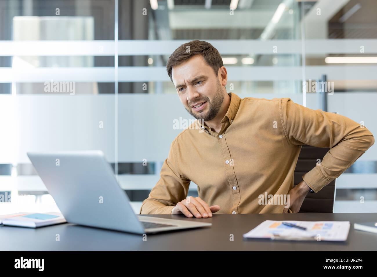 Geschäftsmann sitzt an einem Schreibtisch und arbeitet an einem Laptop, erlebt Unbehagen und Schmerzen im unteren Rücken, betont Probleme durch langes Sitzen und schlechte Haltung Stockfoto