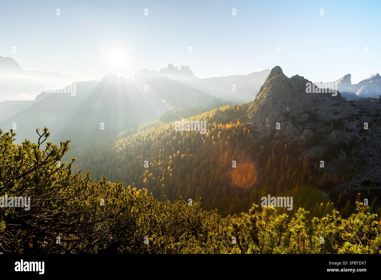 Sonnenstrahlen brechen durch den Bergnebel über Herbstwäldern in den Dolomiten. Atmosphärische Alpenlandschaft mit goldenen Lärchen und geschichteten Bergkämmen Stockfoto