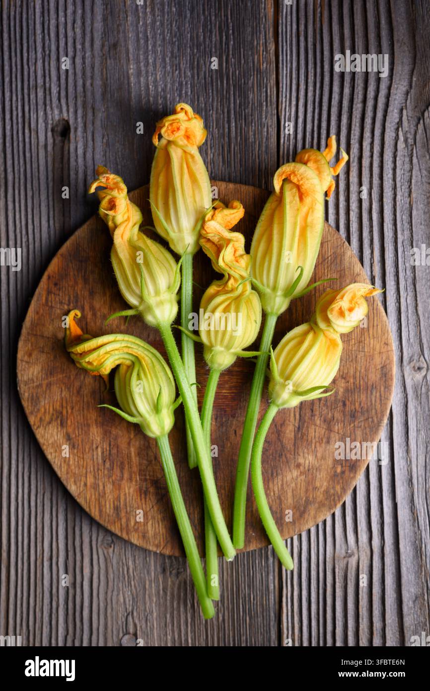 Über Kopf gelegte Zucchiniblüten auf rundem Holzbrett auf rustikaler Oberfläche. Frische essbare Blumen mit natürlicher Komposition und Textur Stockfoto