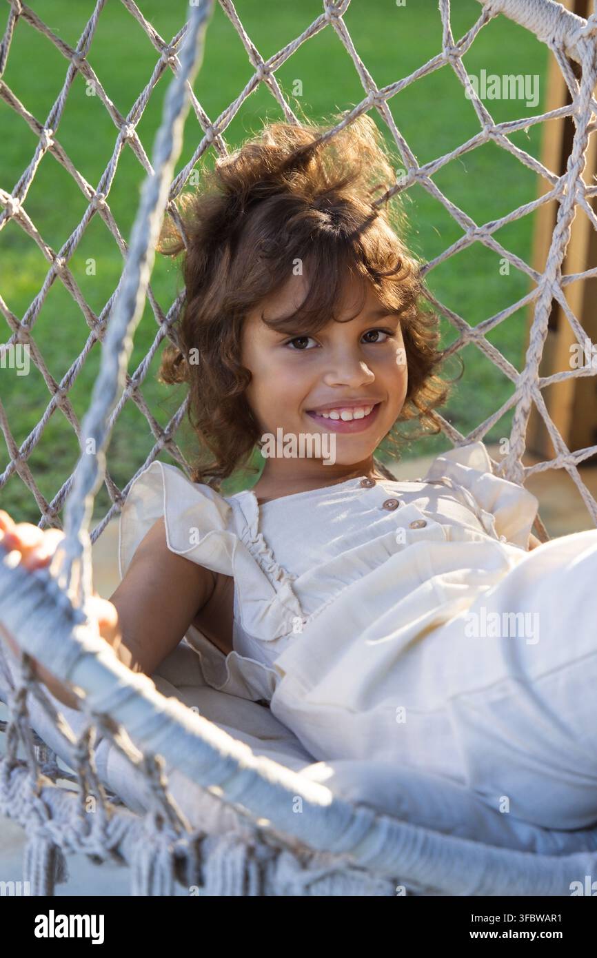 Afroamerikanisches Kind liegt in einem Netzhängematten im Garten mit gewebten Seilen, Holzpfosten, cremefarbenem Kleid Stockfoto