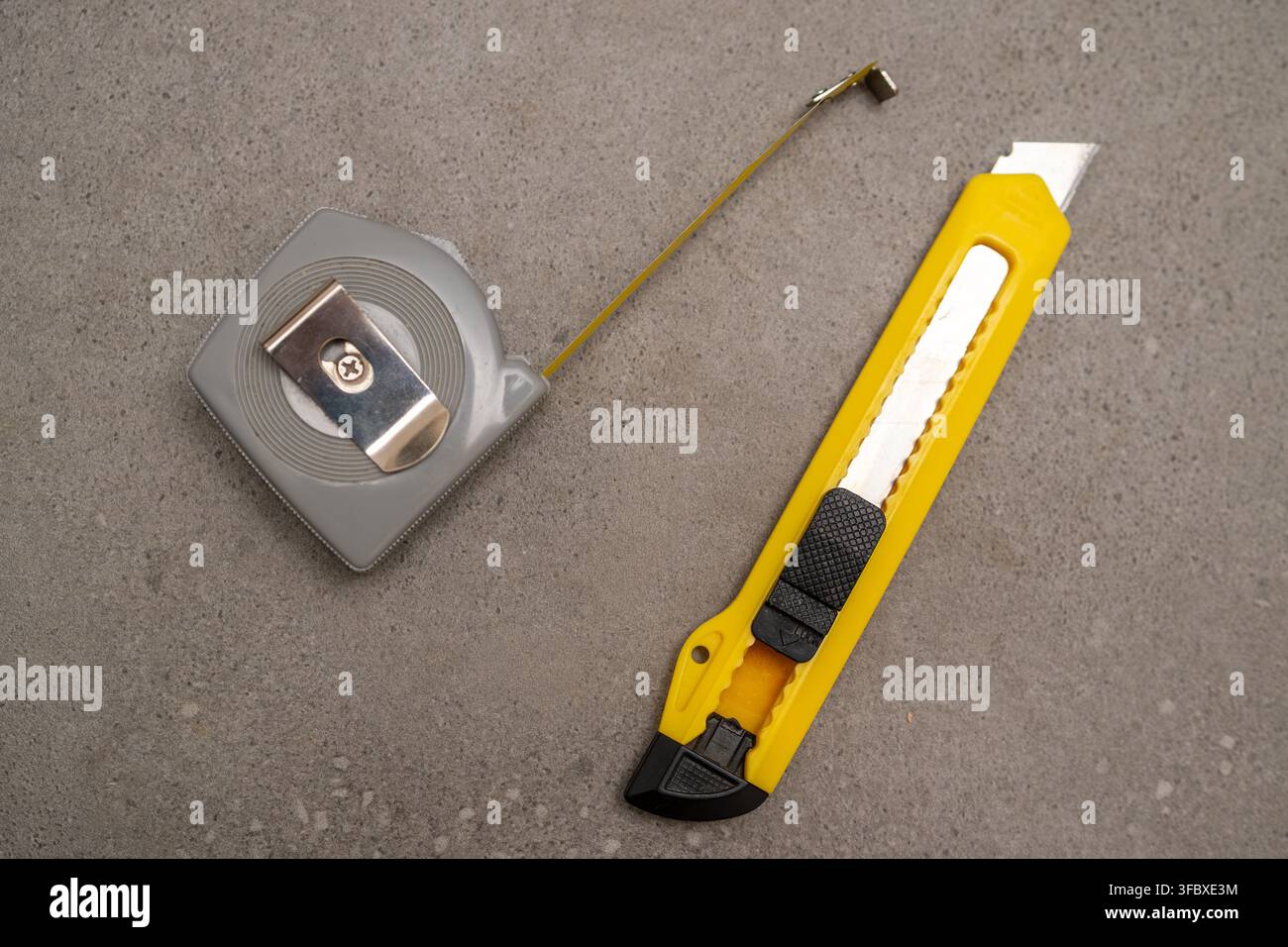 Ein einziehbares Maßband liegt neben einem gelben Universalmesser auf einer Betonoberfläche. Das saubere, minimalistische Layout betont die wesentlichen Werkzeuge, die bei Bau- und Renovierungsprojekten verwendet werden. Stockfoto
