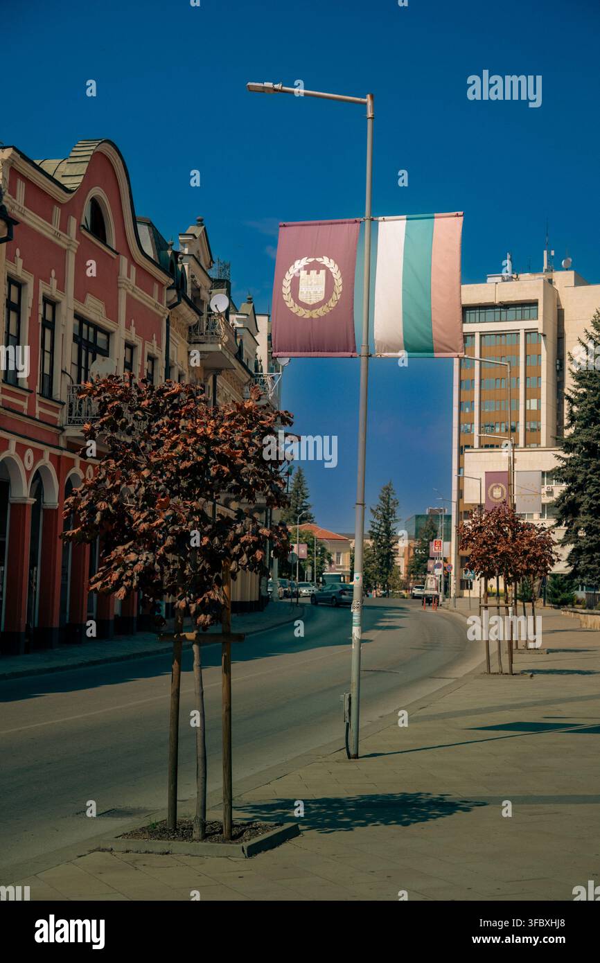 Straße in Lovech mit der regionalen purpurfarbenen Flagge und der bulgarischen Nationalflagge auf einem Mast, mit eleganten architektonischen Gebäuden und einem blauen Himmel. Stockfoto