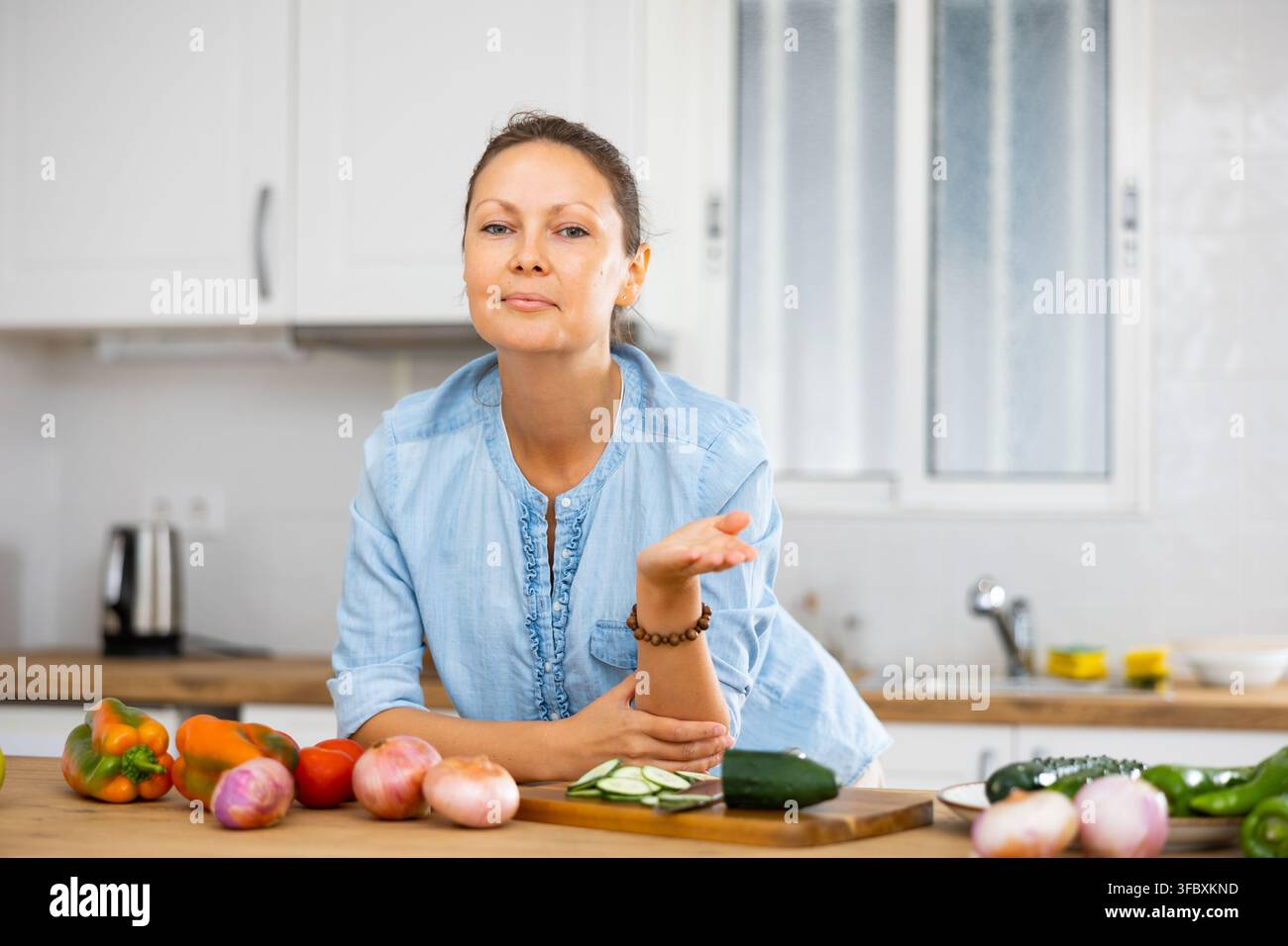 Positive Hausfrau lehnt sich an ein Küchenset mit Gemüse, während sie Salat zubereitete Stockfoto