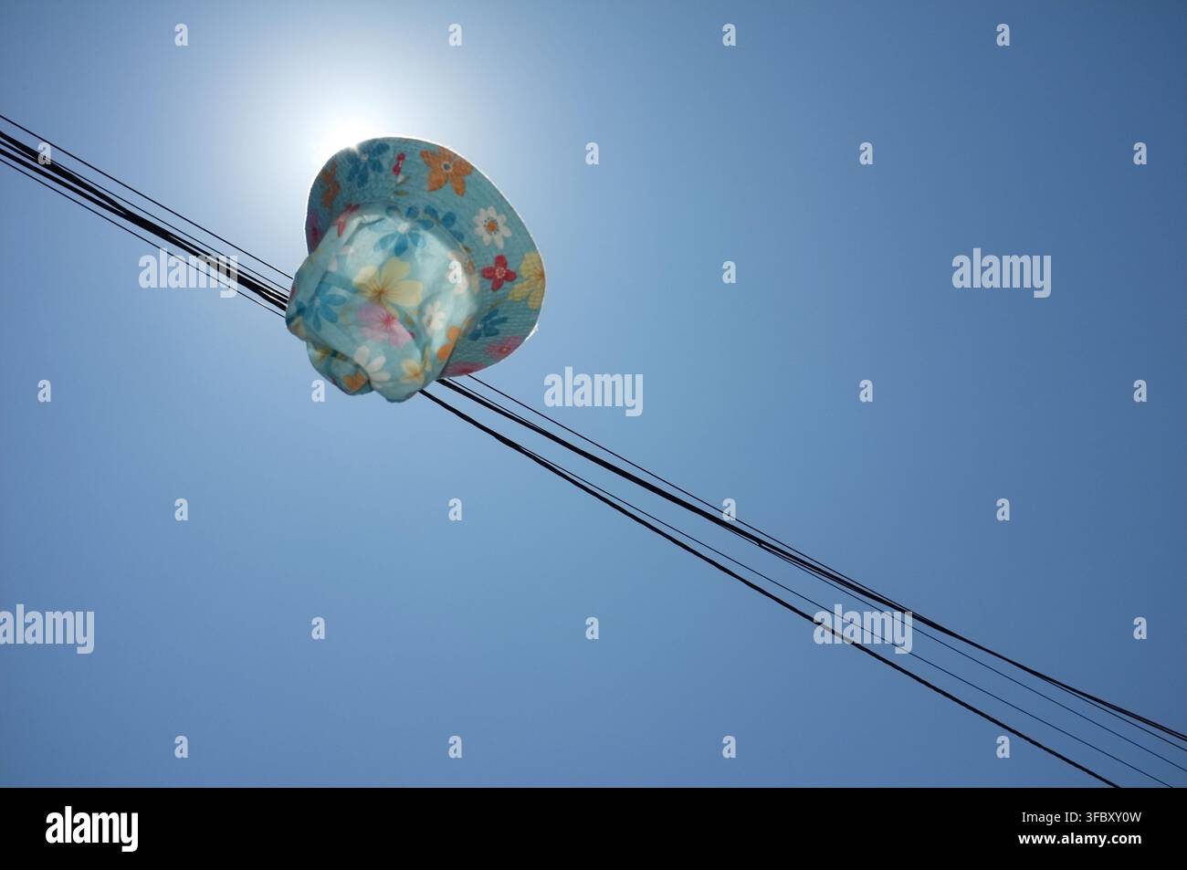 Kindersonnenhut mit Blumenmuster, der gegen einen klaren blauen Himmel in die Luft geworfen wird, beleuchtet von hellem Gegenlicht und eingerahmt von diagonalen Überkopftelefonleitungen. Stockfoto