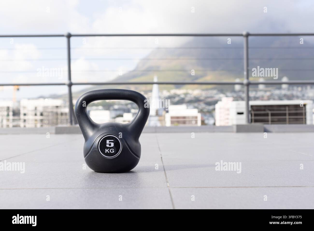 Schwarzer 5-KG-Kettlebell sitzt auf einer gefliesten Dachterrasse, Geländer, Stadtbild und neblige Berge im Hintergrund Stockfoto