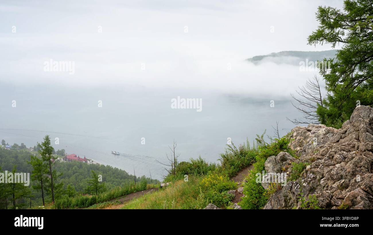 Hochwinkelaufnahme des nebligen Baikalsees von einer felsigen Bergklippe aus, Sibirien. Kleines Boot navigiert nahe einem bewaldeten Ufer. Atmosphärische Naturlandschaft Stockfoto