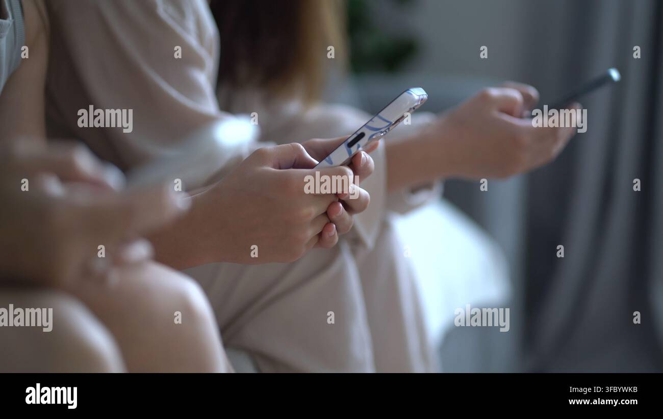 Nahaufnahme von jungen Frauen, die drinnen im weichen natürlichen Licht auf Smartphones scrollen, lässige beige Kleidung, moderner Lebensstil-Moment des Digitalen Stockfoto