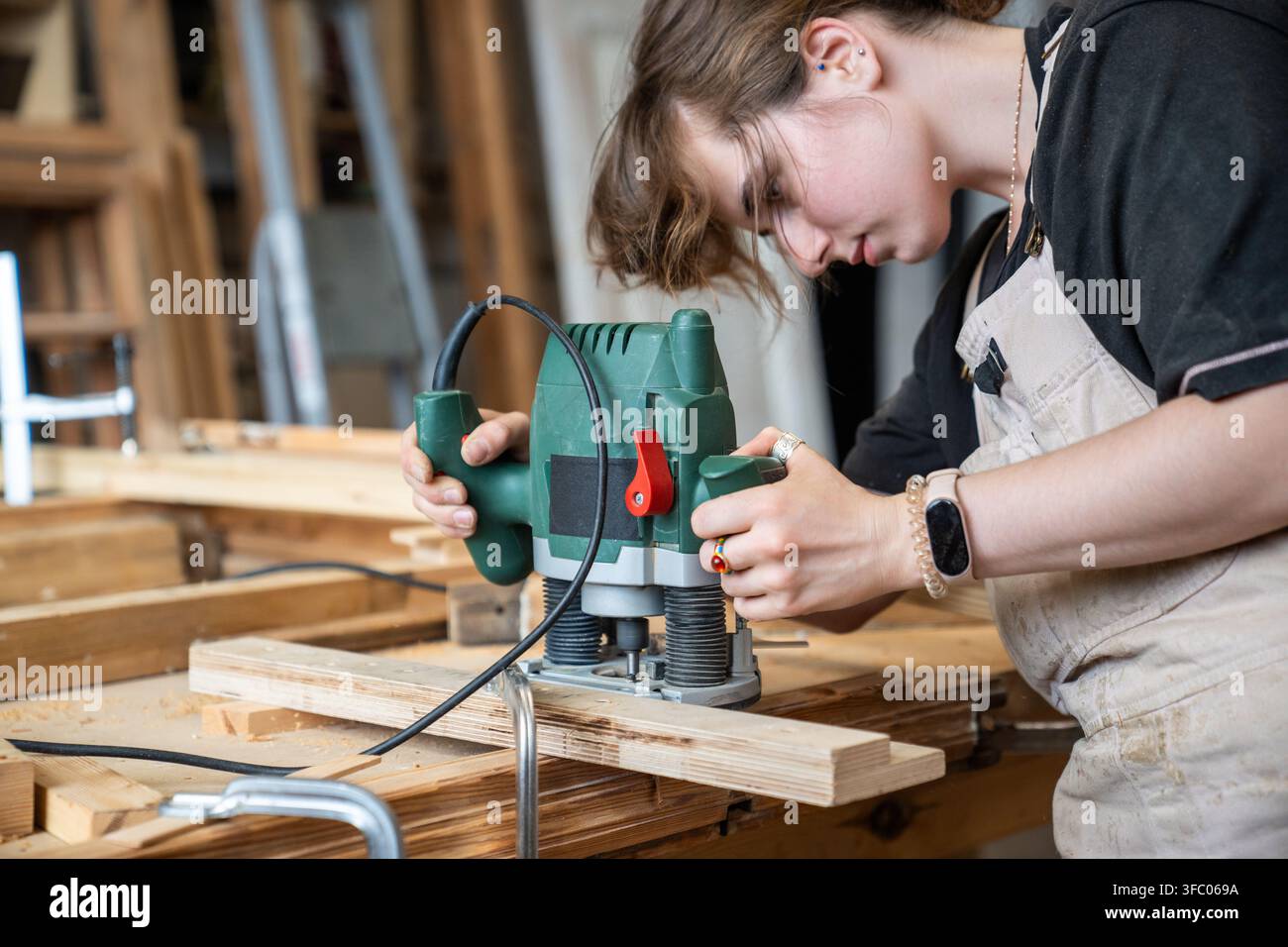 Konzentrierte, geschickte Restauratorin arbeitet mit einer Elektrotauchfräse und schneidet eine Oberfläche an einem Holzdetail Stockfoto