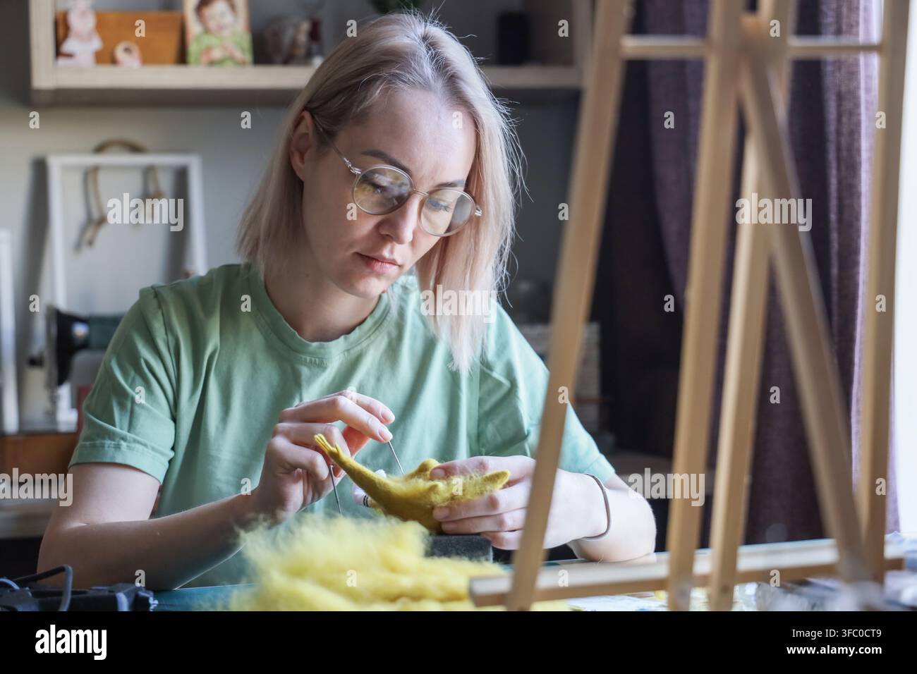 Junge Frau mit Brille stickt sorgfältig eine gelbe Wollfigur an einem Basteltisch zu Hause, arbeitet mit konzentriertem Ausdruck im sanften natürlichen Tageslicht Stockfoto