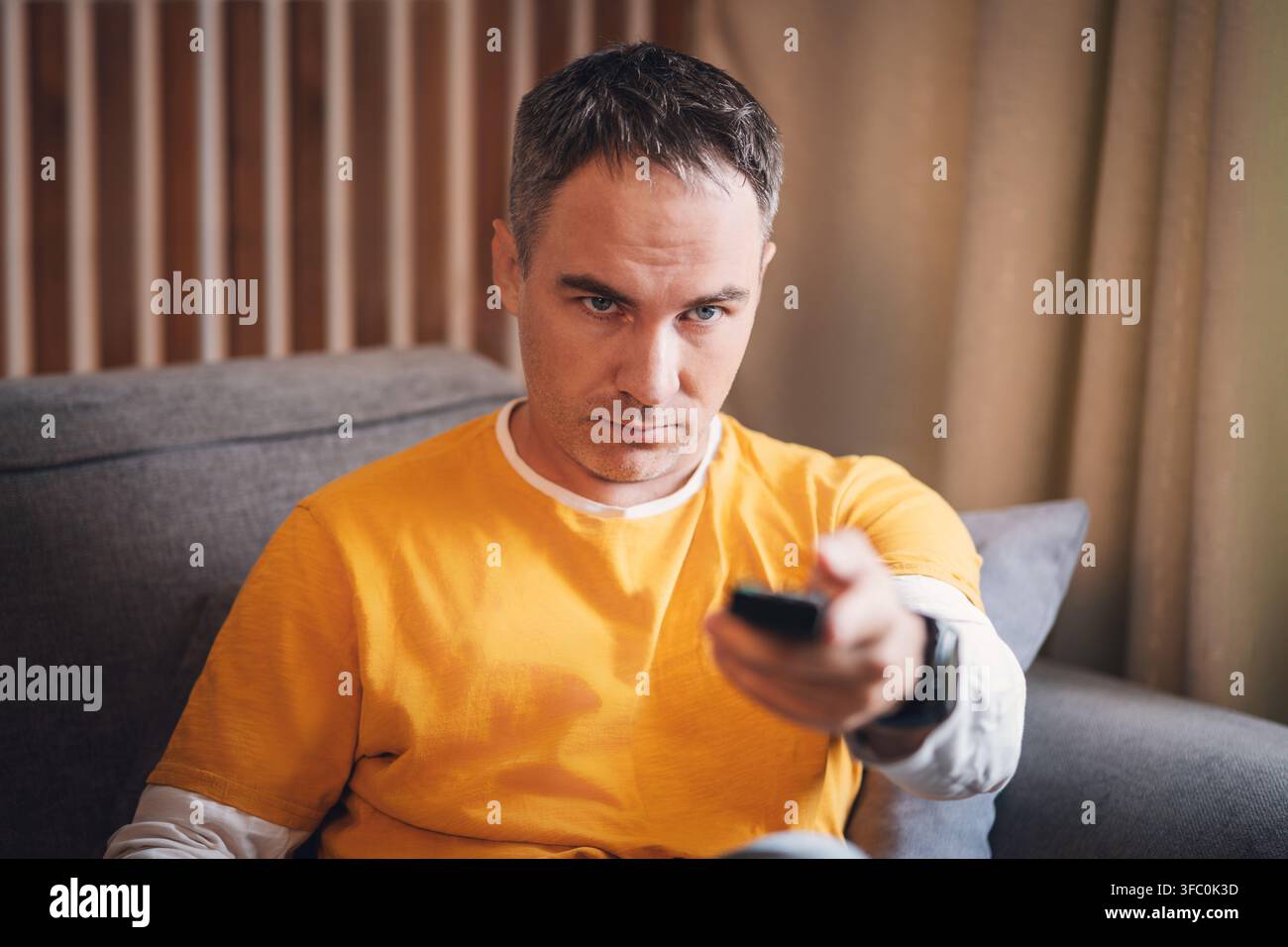 Mittelalter Mann in einem senfgelben T-Shirt sitzt auf einem grauen Sofa und hält eine TV-Fernbedienung; konzentrierter und ernster Ausdruck. Holzlatten und beigefarbene Vorhänge i Stockfoto