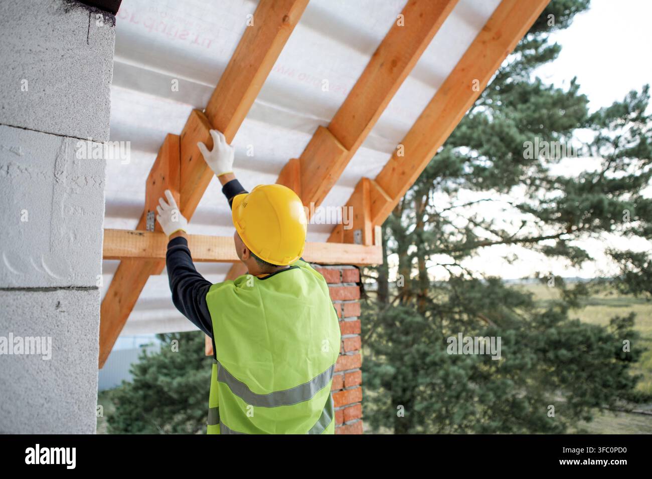 Bauarbeiter sichert Dachsparren mit Werkzeug, Haus im Bau mit sichtbaren Holzsparren, Zusammenbau des Hauptdachgerüsts Stockfoto