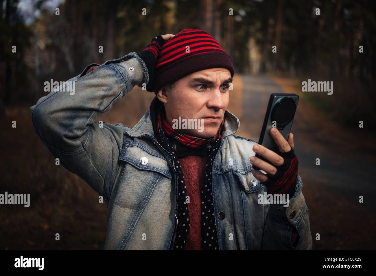 Mann hält sich den Kopf in Schock, während er auf sein Smartphone in einer Waldkulisse schaut, sich verloren fühlend. Kein Signal, keine Verbindung und gestrandet im Wald Stockfoto