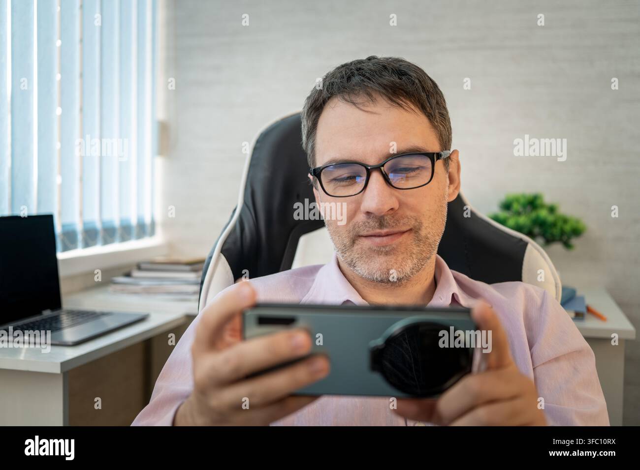 Lächelnder Mann mit Brille, der ein Smartphone im Querformat benutzt, während er im Büro entspannt und mobile Unterhaltung genießt. Stockfoto