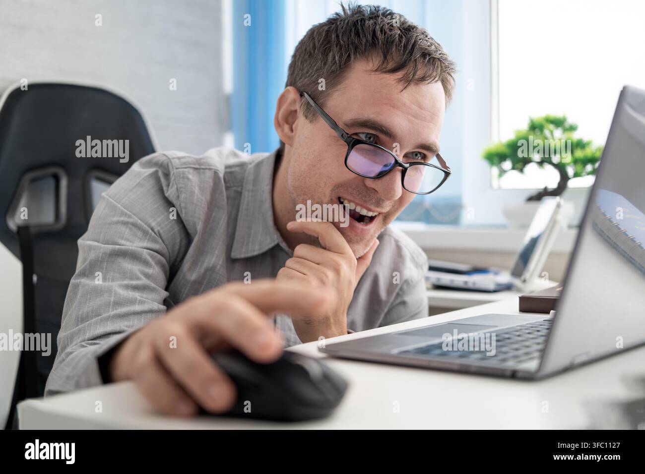 Zufriedener Mitarbeiter lächelt, während er einen Laptop und eine Computermaus im Büro verwendet, und genießt erfolgreiche Online-Arbeit, positive Ergebnisse und produktives digitales Kommunizieren Stockfoto