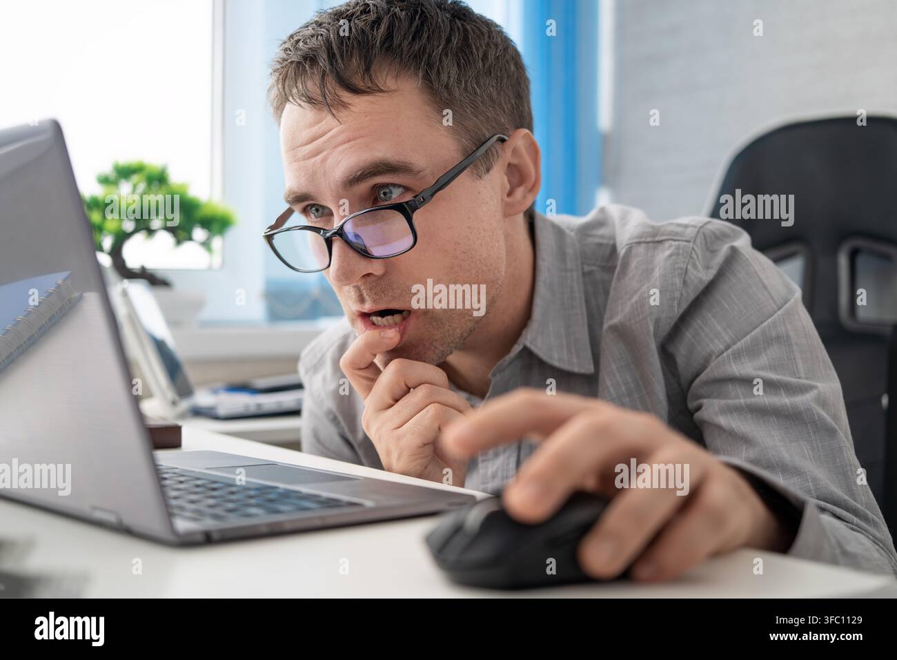 Ein überraschter Büroangestellter in einem grauen Hemd und einer Brille reagiert auf etwas auf dem Laptop-Bildschirm; er hält mit einer Hand eine Maus und berührt mit der anderen sein Kinn Stockfoto