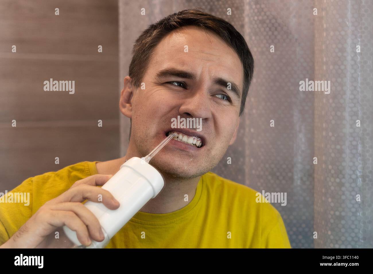 Mann, der vor Schmerzen das Gesicht verzieht, während er einen Wasserflosser an empfindlichem Zahnfleisch verwendet. Zahnschmerzen, Zahnfleischsensibilität und unangenehme Mundpflege. Stockfoto