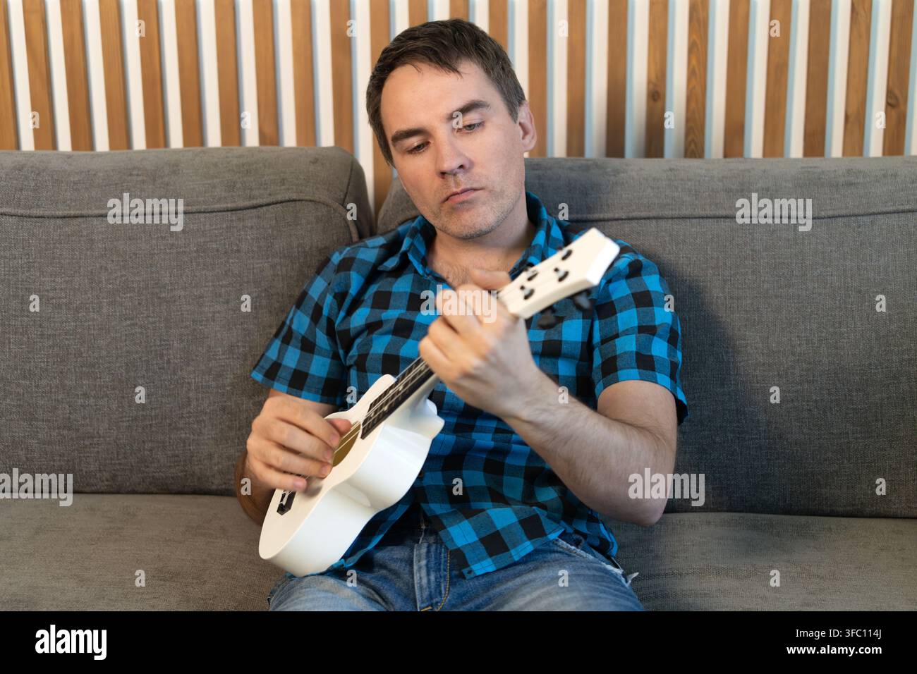 Mann sitzt auf dem Sofa und lernt Ukulele spielen, musikalisches Hobby und Heimübung. Stockfoto