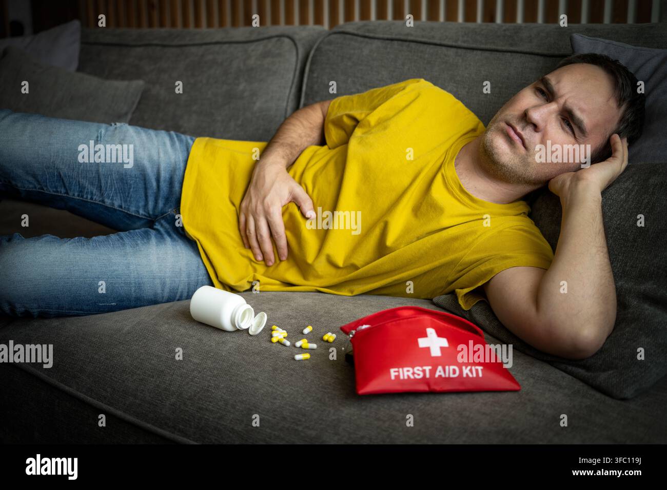 Kranker Mann liegt auf dem Sofa und hält sich den Bauch, verstreute Pillen und ein Erste-Hilfe-Koffer in der Nähe, gesundheitlicher Notfall. Stockfoto