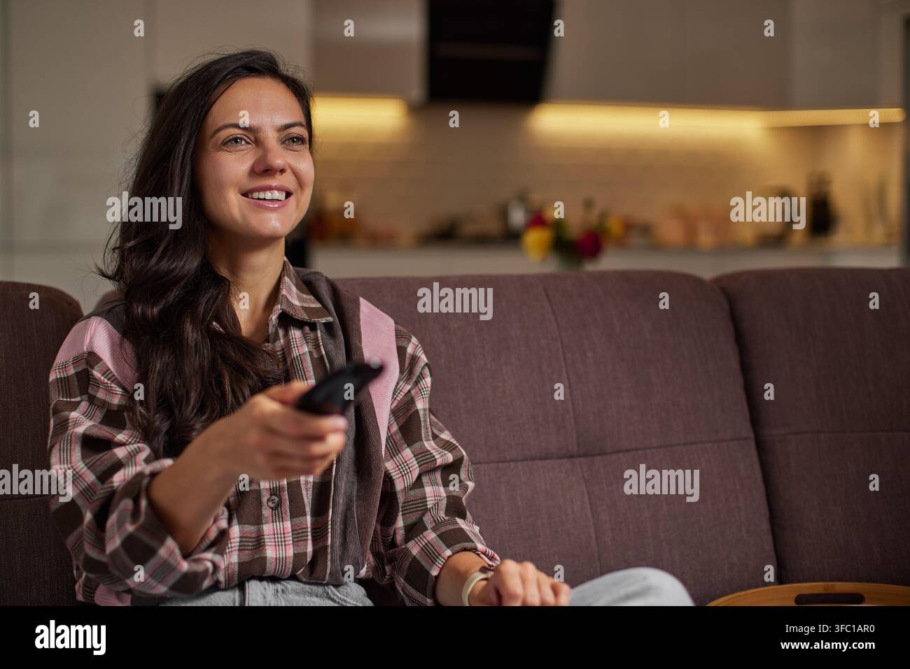 Lächelnde Frau sitzt auf einer Couch und hält eine Fernbedienung. Sie schaut auf einen Fernsehbildschirm und genießt ihre Freizeit zu Hause Stockfoto