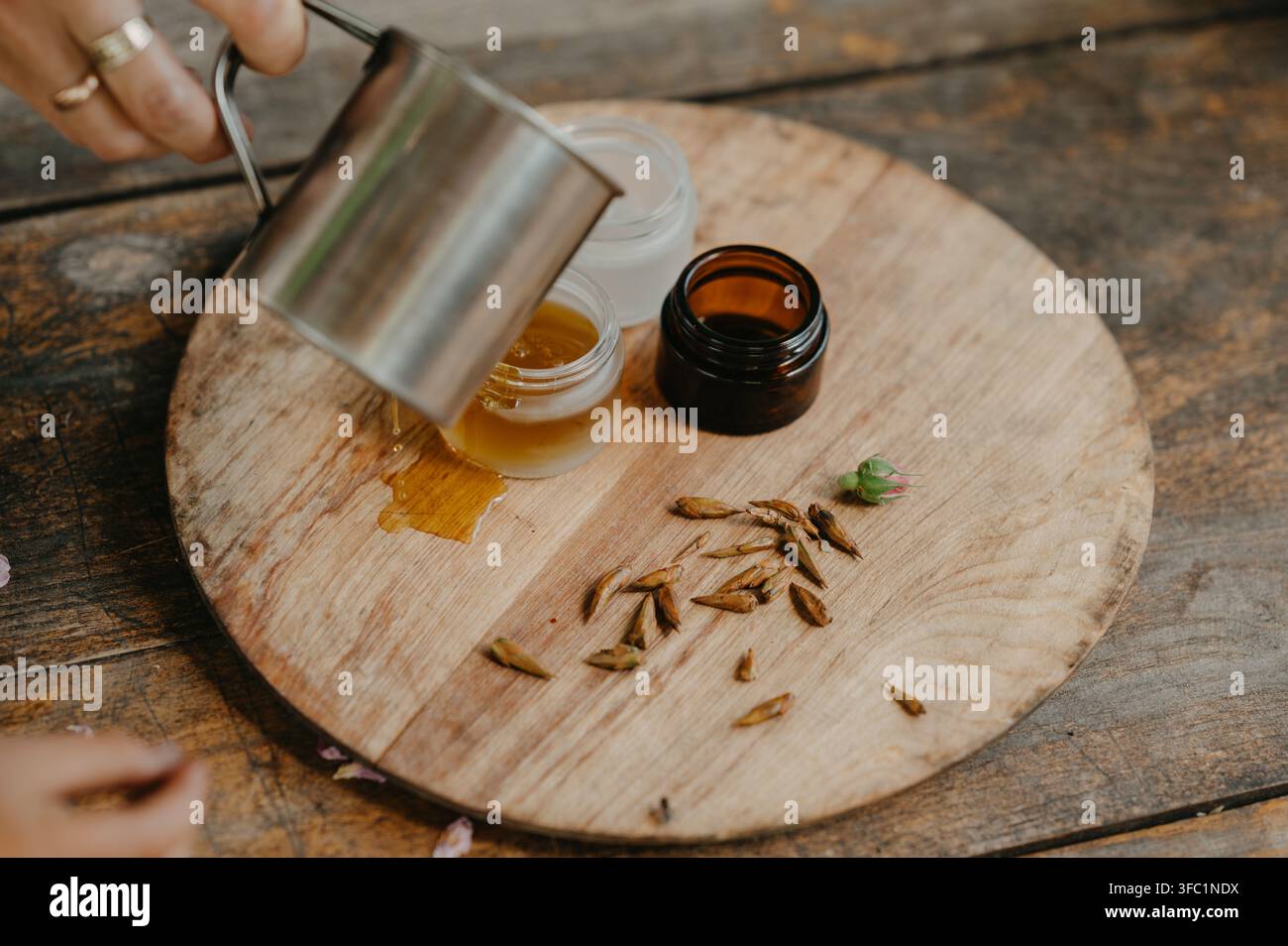 Hand gießt goldene Flüssigkeit in ein kleines Glasgefäß auf einer rustikalen Holzplatte, mit verschütteten Tropfen, getrockneten Samenschalen, einem bernsteinfarbenen Behälter und natürlichem handgefertigtem Pr Stockfoto