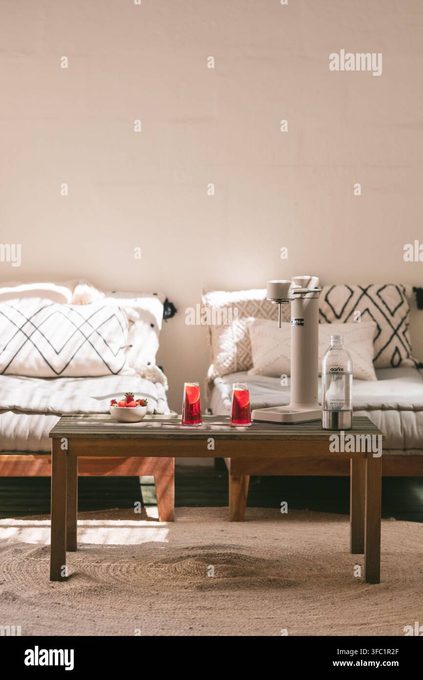 Helle Boho-Wohnzimmer mit einem Holzkaffeetisch, roten Cocktails, einem Sodamixer, Sofa mit Kissen und einer Pflanze, gemütliche Hausaperitif-Lebensstil Stockfoto