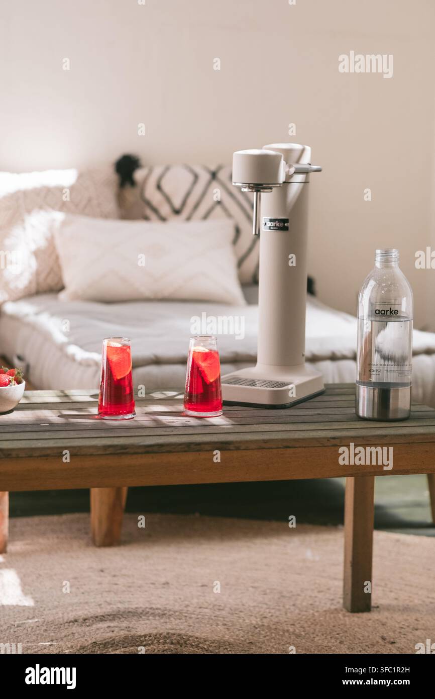 Zwei rote Cocktails in hohen Gläsern neben einem Sodamixer und einer Flasche auf einem hölzernen Couchtisch in einem hellen Wohnzimmer, hausgemachte Getränke zu Hause Stockfoto
