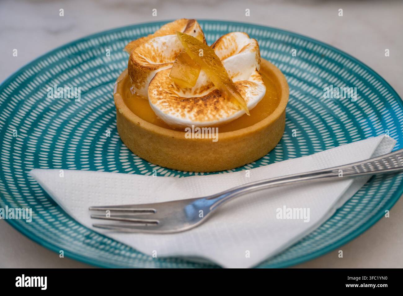 Eine frisch gebackene Mini-Zitronen-Baiser-Tarte, garniert mit kandierter Schale, liegt auf einem blau gemusterten Keramikteller neben einer weißen Serviette und einem silbernen Dessertlöffel Stockfoto