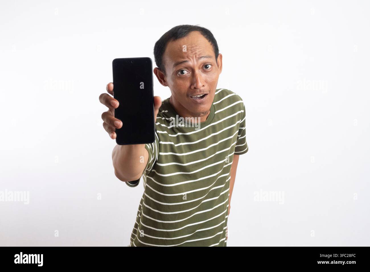 Ein asiatischer Mann in einem grünen Hemd lächelt, während er ein Smartphone mit einem leeren Bildschirm hochhält. Der Hintergrund ist einfarbig weiß, und der Mann zeigt auf die Ka Stockfoto