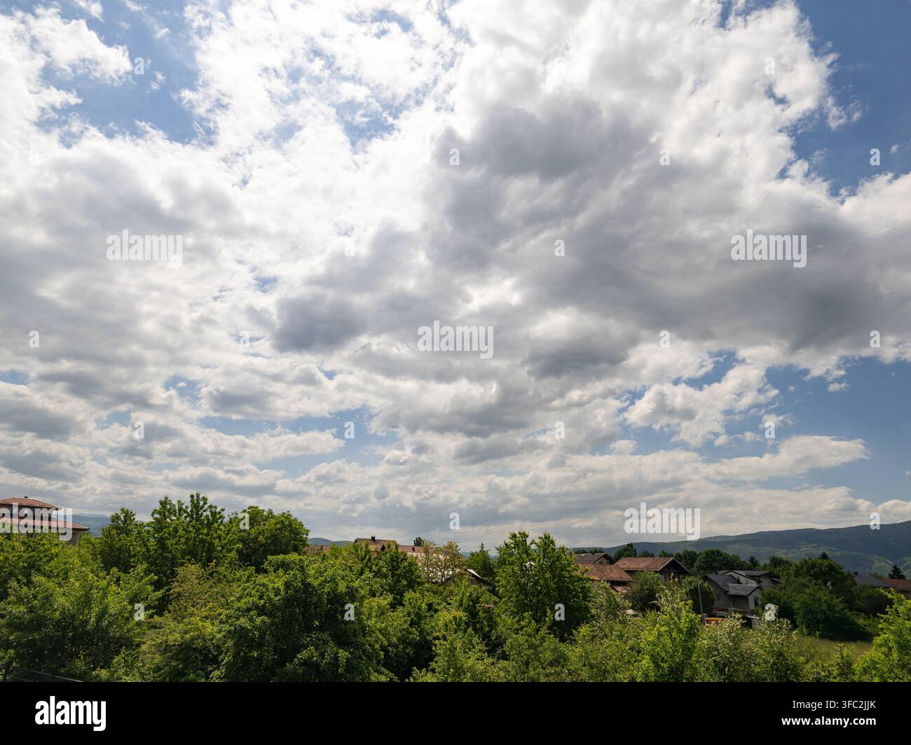 Ein malerischer Blick auf ein ruhiges Dorf mit Häusern, die sich zwischen üppigen grünen Bäumen verstecken, unter einem weiten, dramatischen, wolkenverhangenen Himmel. - Smartphone-aufgenommenes Stockfoto