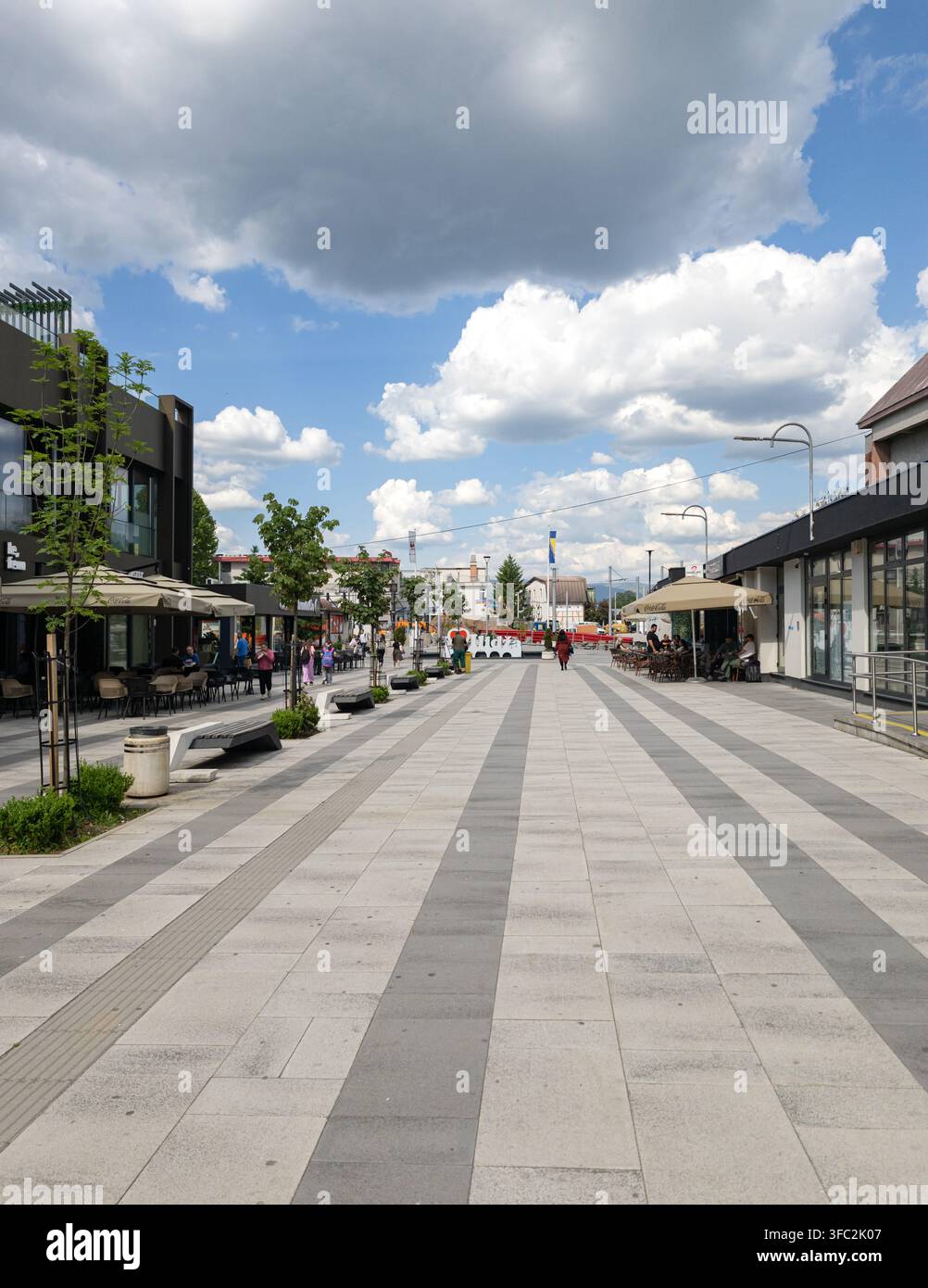 Fußgänger überqueren einen breiten, gepflasterten modernen Stadtplatz in Ilidza, Bosnien, unter einem strahlenden Himmel mit dramatischen Kumuluswolken. - Smartphone-aufgenommenes Stockfoto
