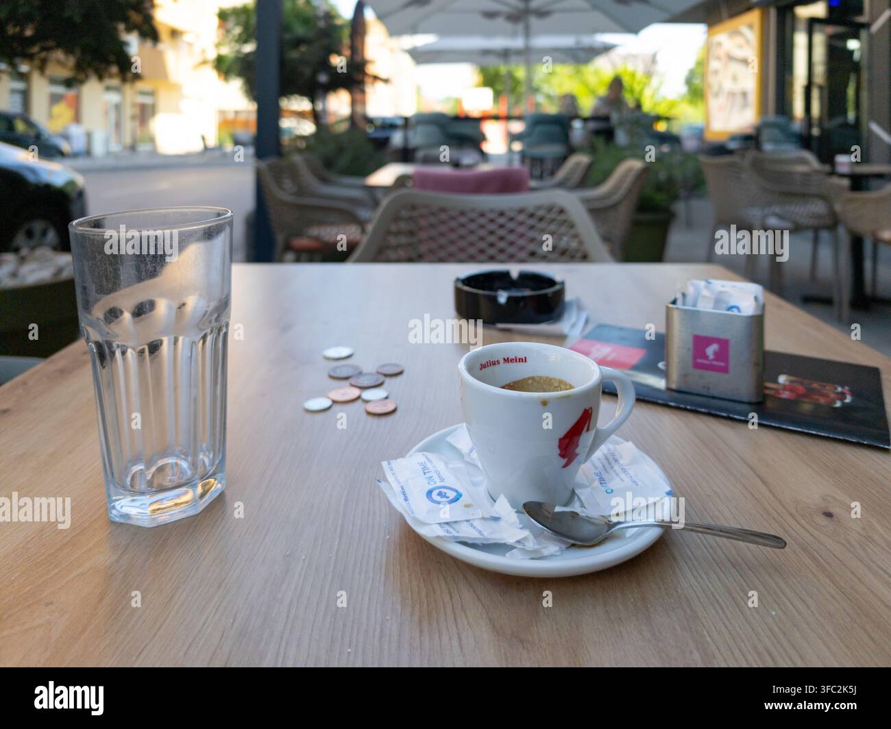 Eine Tasse Julius Meinl Kaffee, ein leeres Glas und lose Münzen, die auf einem Außentisch des Cafés während einer friedlichen Nachmittagspause ruhen. - Smartphone-aufgenommenes Stockfoto