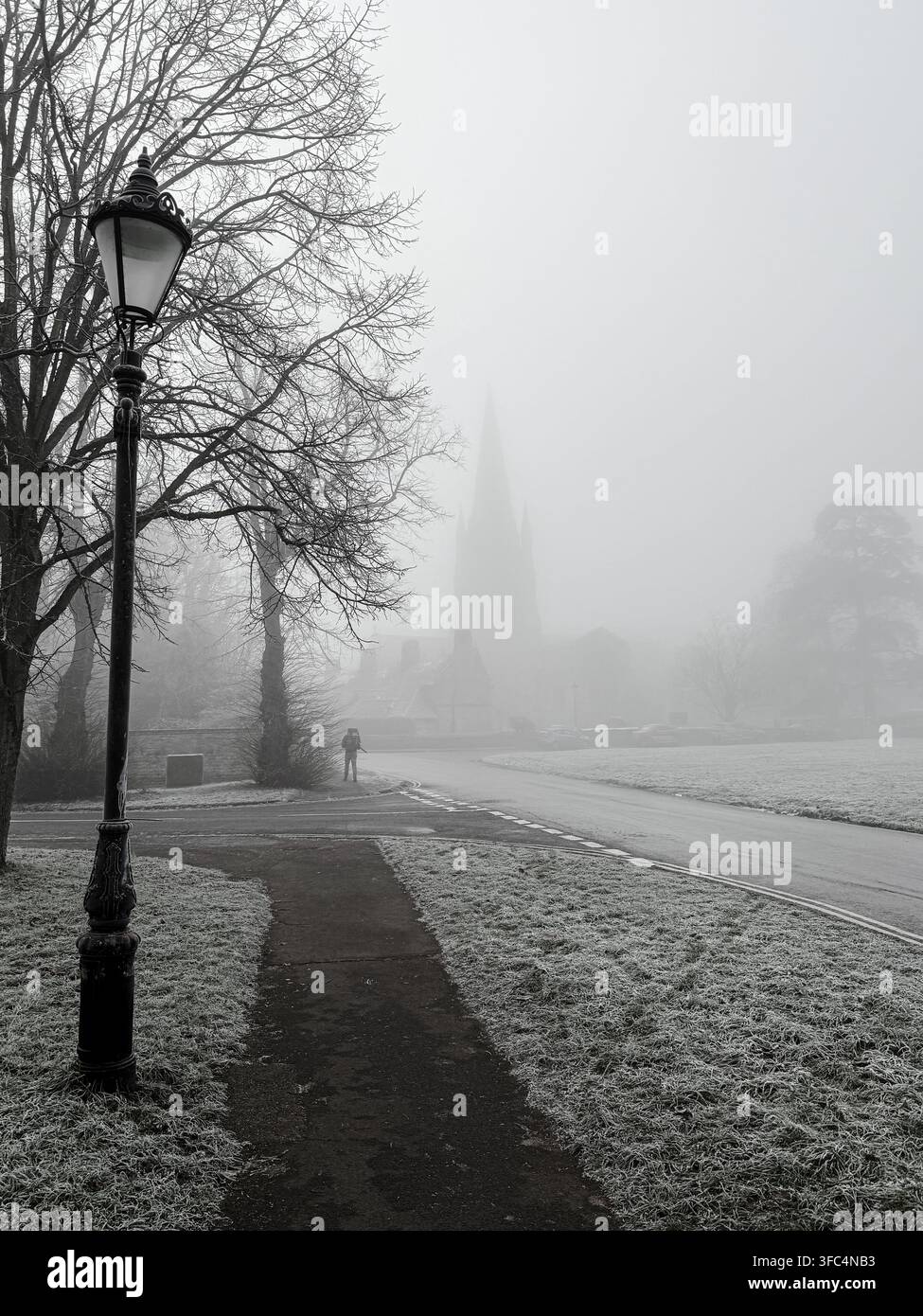 Dichter Nebel hüllt eine ruhige Dorfstraße ein, während eine einsame Gestalt an einer Laternenpfähle vorbeigeht in Richtung eines Kirchturms, der im Nebel verschwindet. Stockfoto