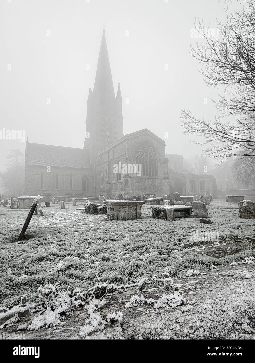 Frost bedeckt Grabsteine in einem historischen Friedhof, während sich der Turm einer gotischen Kirche in dichten Morgennebel erhebt. Stockfoto