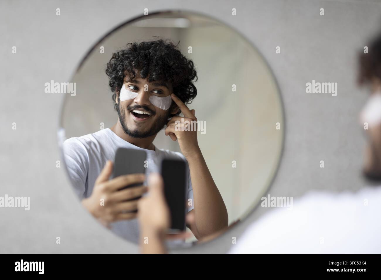 Glücklicher junger Mann, der vor dem Spiegel zu Hause eine Gesichtsmaske aufträgt, Hautpflegeroutine Stockfoto