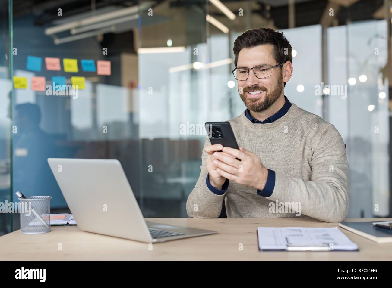 Junger lächelnder Mann mit Bart und Brille sitzt an einem Schreibtisch, schaut auf sein Mobiltelefon, während er in einer zeitgenössischen Unternehmensumgebung mit einem Laptop arbeitet Stockfoto