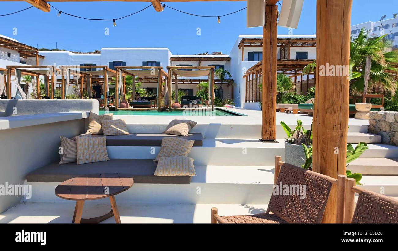 MYKONOS, GRIECHENLAND - 15. JULI 2026: Ein ruhiger Außenlounge-Bereich mit bequemen Kissen, Holzsitzen und weißen mediterranen Gebäuden an einem Stockfoto