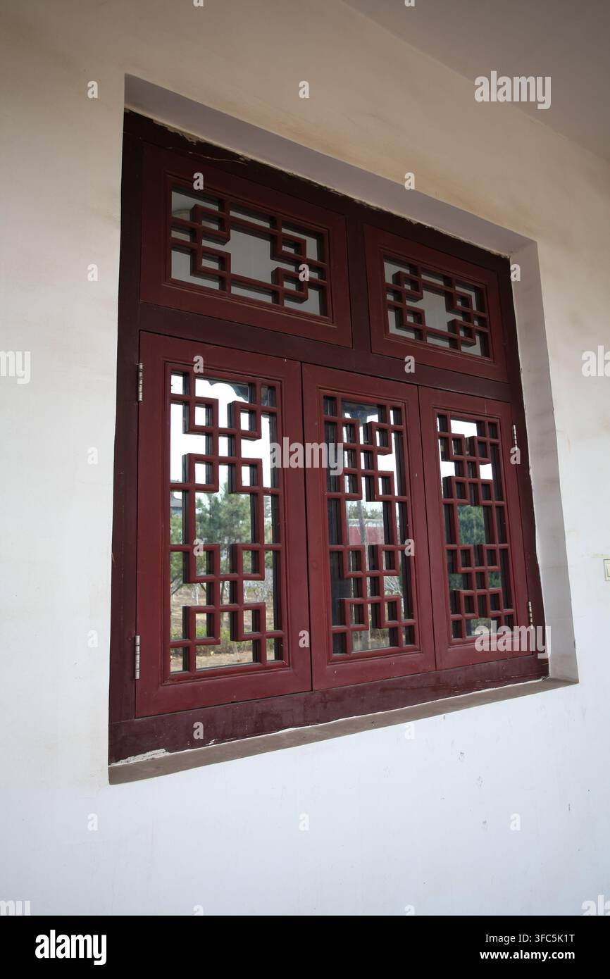 Ein rötlich-braunes Holzfenster im chinesischen Stil an einer weißen Wand, das traditionelle geometrische Gitterarbeiten mit sauberen und symmetrischen Linien aufweist, ein typi Stockfoto