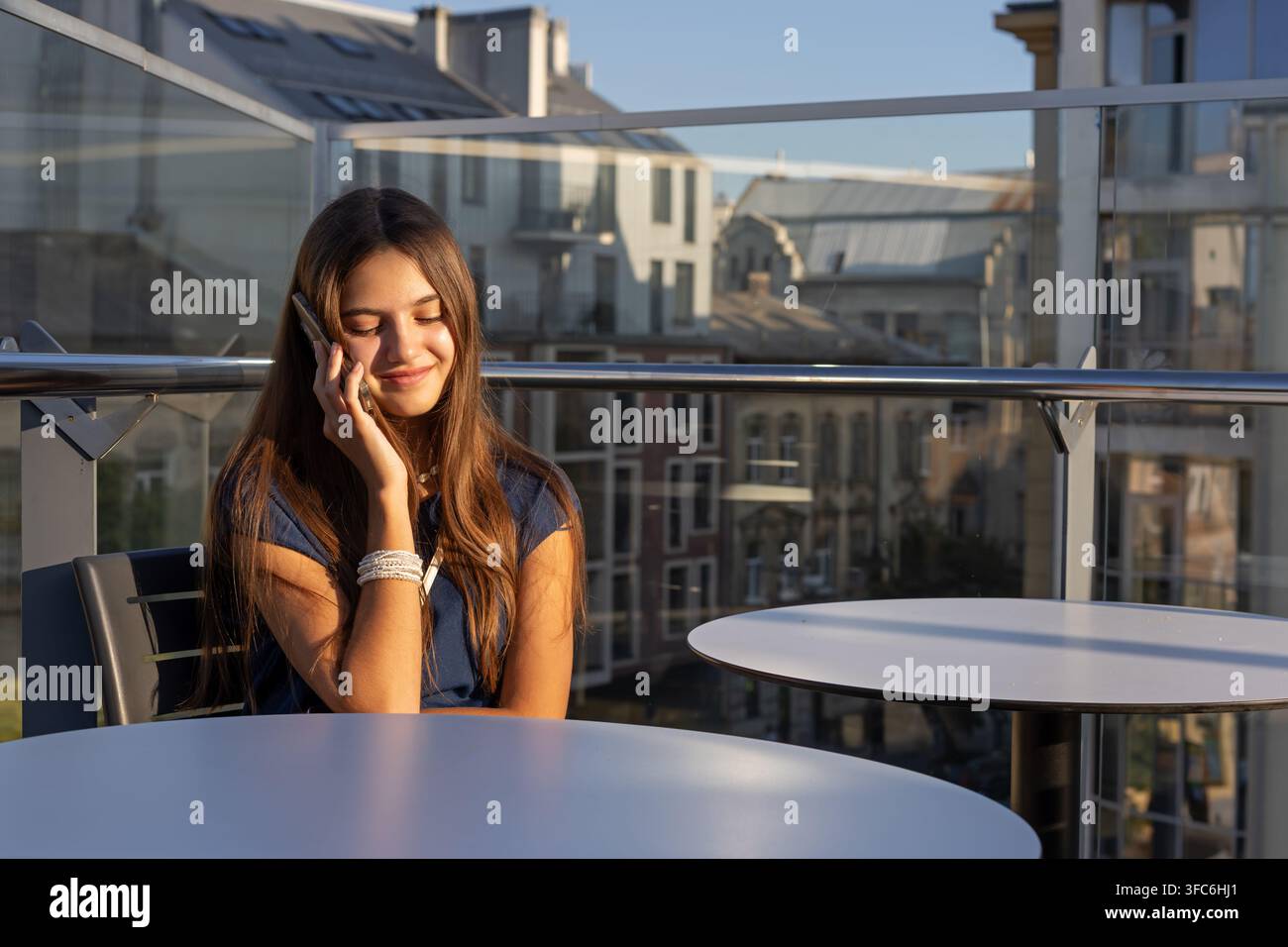 Junge Frau telefoniert auf Dachterrasse. Junge Frau lächelt, während sie an einem Smartphone auf einem Tisch der Dachterrasse in warmem Sonnenlicht telefoniert mit Stockfoto
