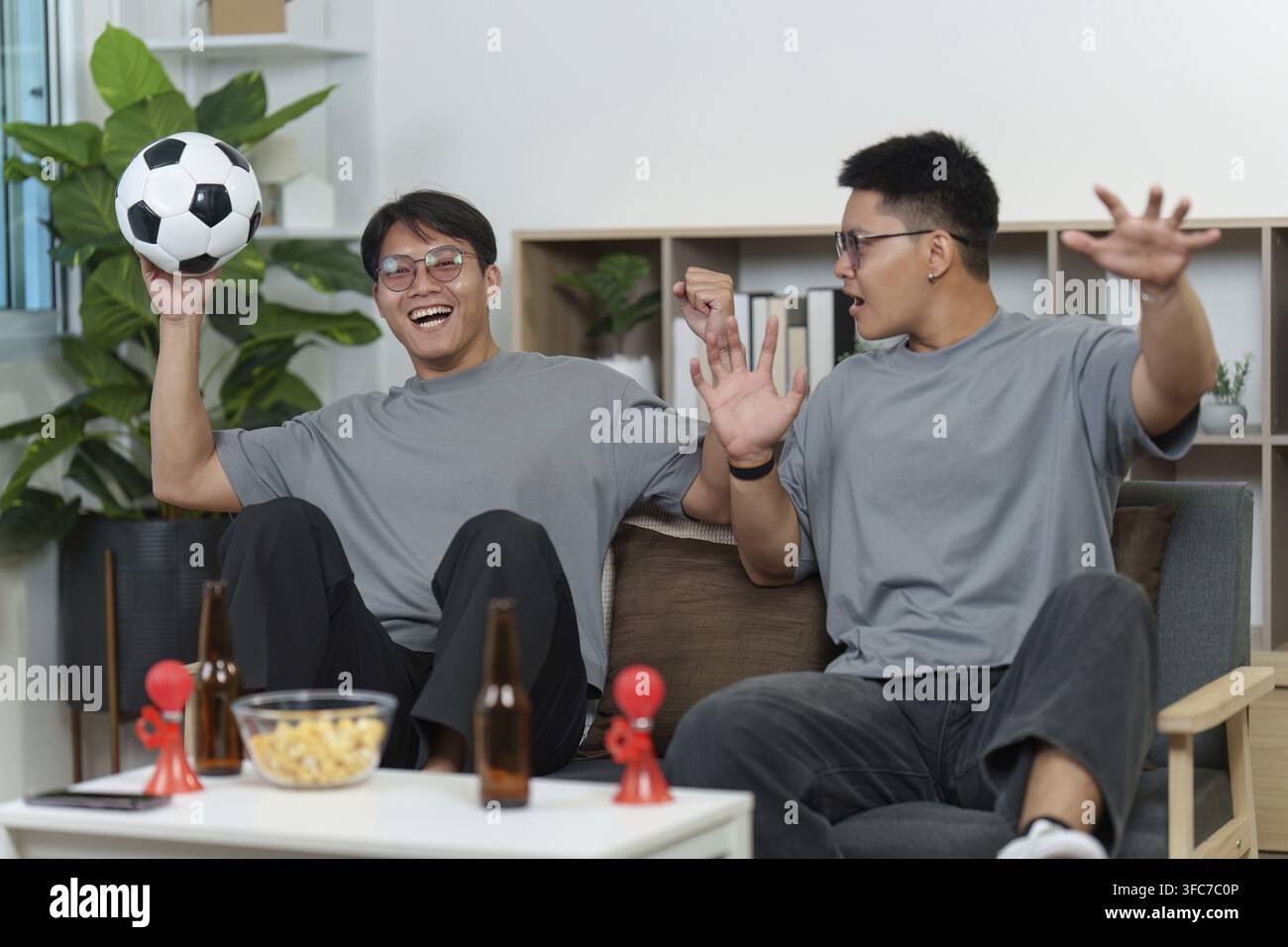 Zwei aufgeregte asiatische Männer, leidenschaftliche Fußballfans, feiern ein Tor mit einem Fußball in den Händen, während sie zusammen in ihrem Wohnzimmer ein Spiel ansehen Stockfoto